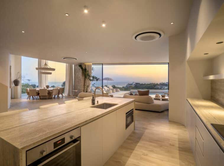 Kitchen view from villa Namiko overlooking the living room and its open windows where the pool, ocean view and a colourful sunset is seen